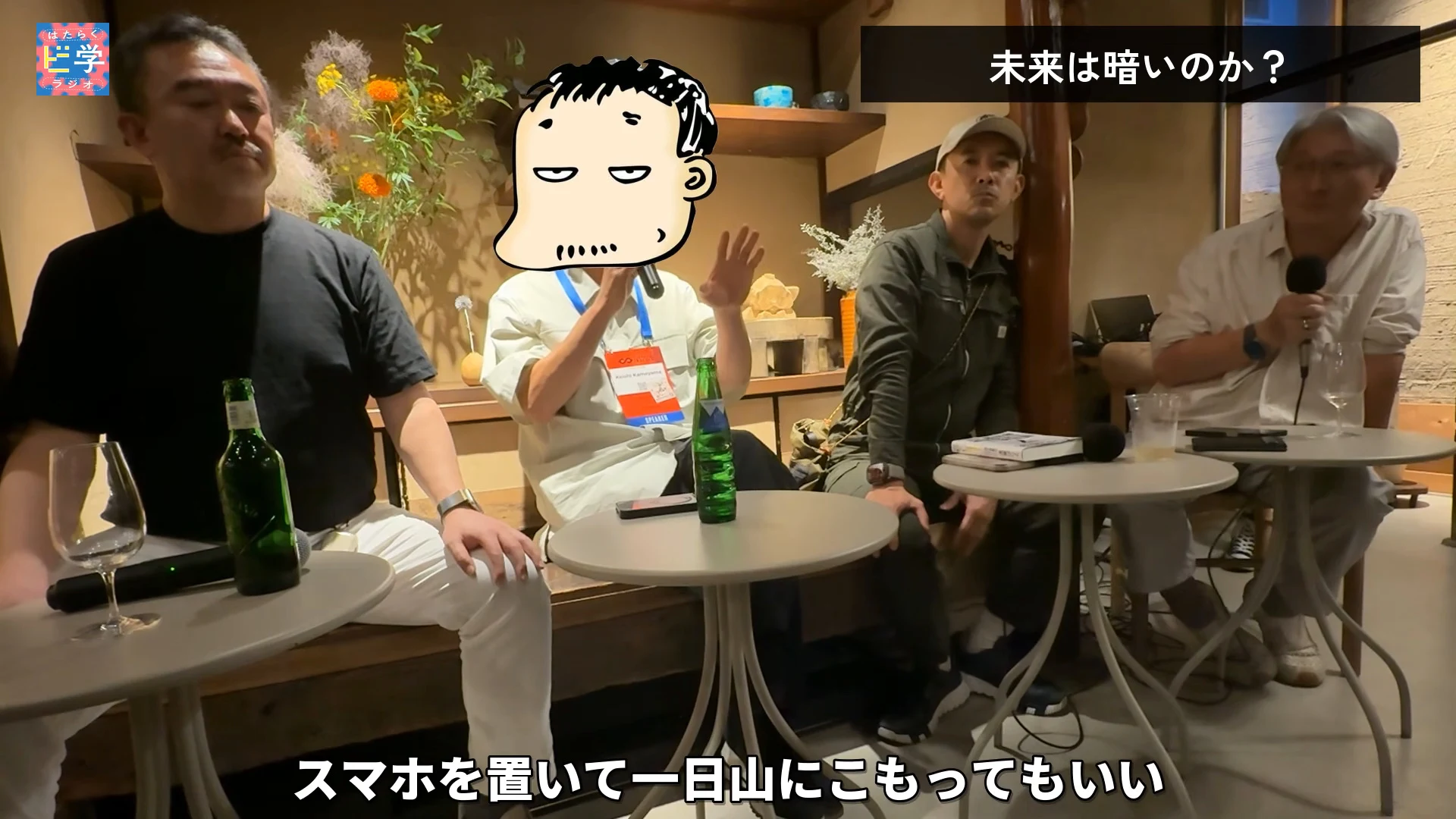 動画の画面。大きく映る黒い服の人、手を挙げて話す司会、右奥の白髪の人。右上のテロップは未来は暗いのか、下のテロップはスマホを置いて一日山にこもってもいいという内容