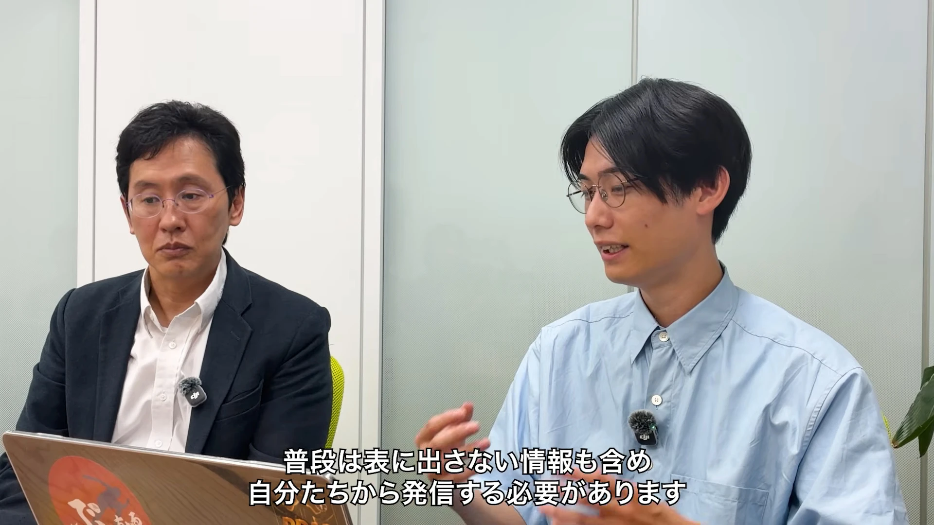 対談動画の場面。右の青いシャツの人が手を動かして話し、左のスーツの人が聞いている