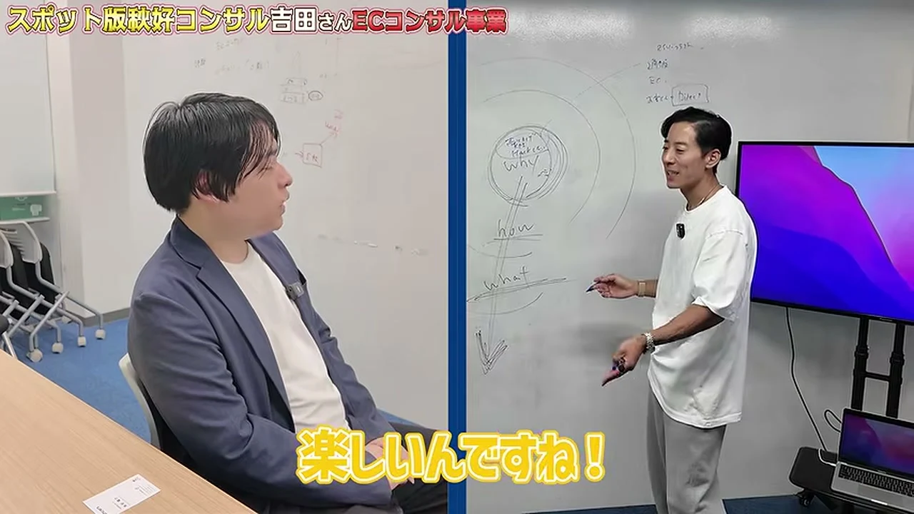 動画の画面。左に椅子に座る紺のジャケットの男性、右のホワイトボードの前に白いTシャツの男性が立っている。字幕に、楽しいんですね、と出ている場面