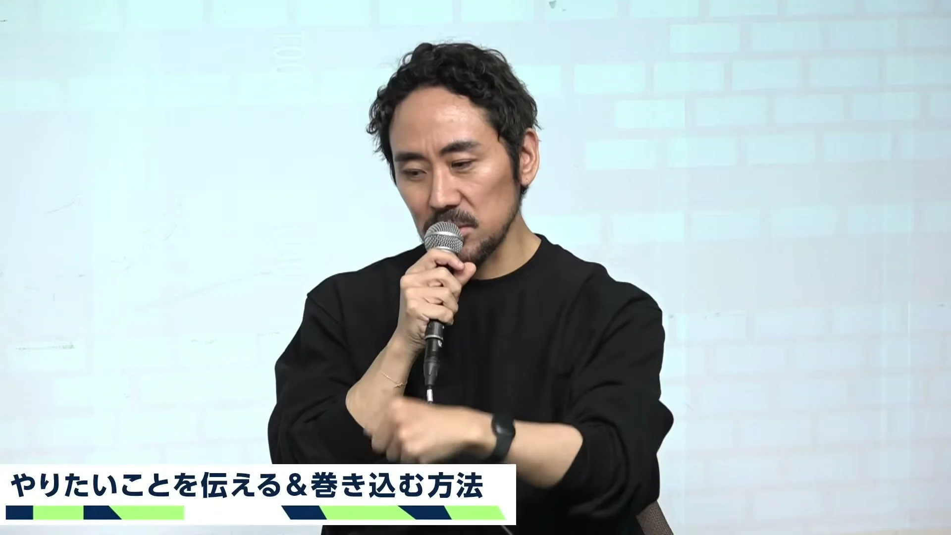 動画キャプチャ: テーマ3、やりたいことを伝える＆巻き込む方法のテロップの下で、マイクを持って話す家入さん