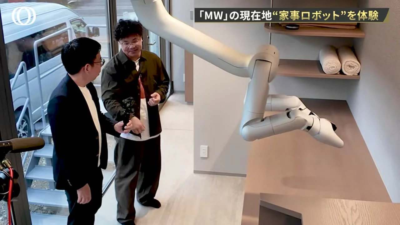 動画の画面。天井から伸びた白いロボットアームが、タオルの並ぶ棚の前の作業台に腕を伸ばし、手前で二人の男性が見ている場面
