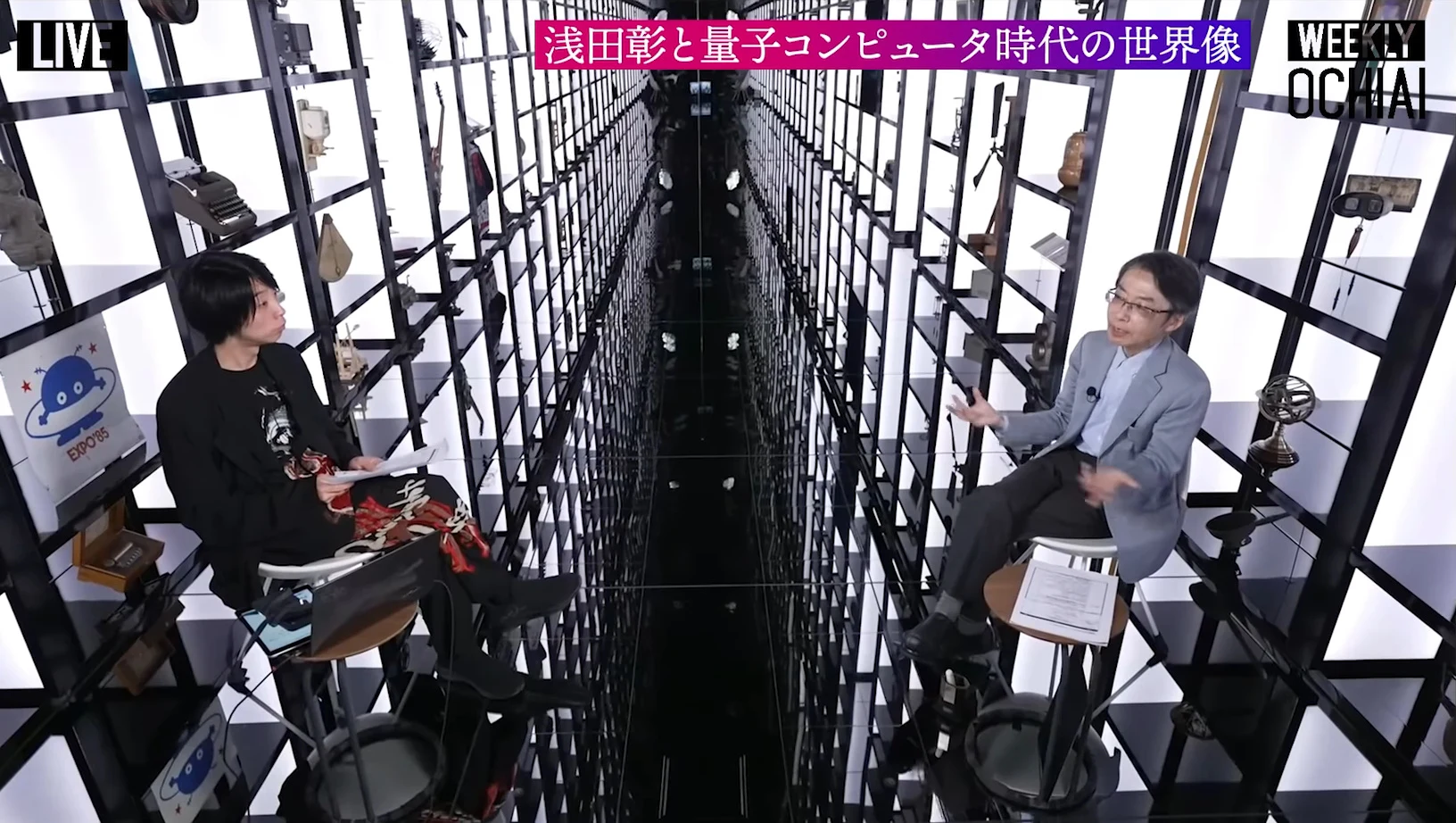 画面上に浅田彰と量子コンピュータ時代の世界像の帯が出て、両側に棚が並ぶ細長い展示室を斜め上から映し、左に黒い服の人物、右に青灰色のジャケットの人物が向かい合って座る動画の場面