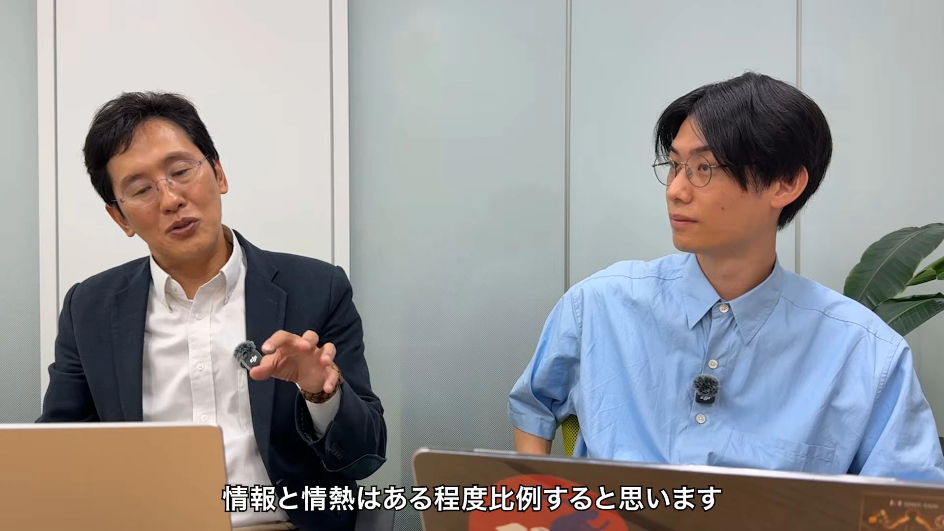 対談動画の場面。左のスーツの人が手を動かして話し、右の青いシャツの人が横を向いて聞いている