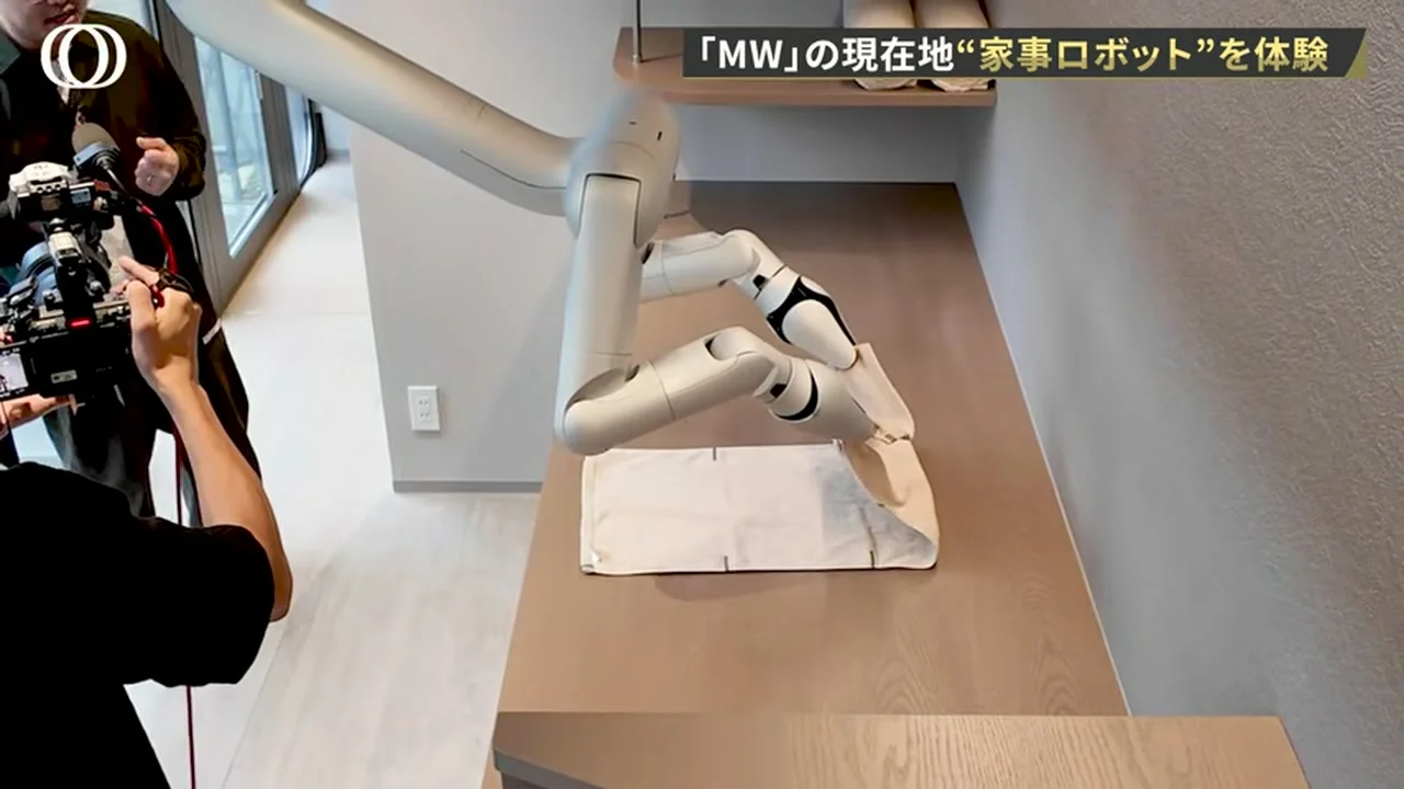 動画の画面。天井から伸びたロボットアームが、作業台の上で白い布を畳んでいる場面