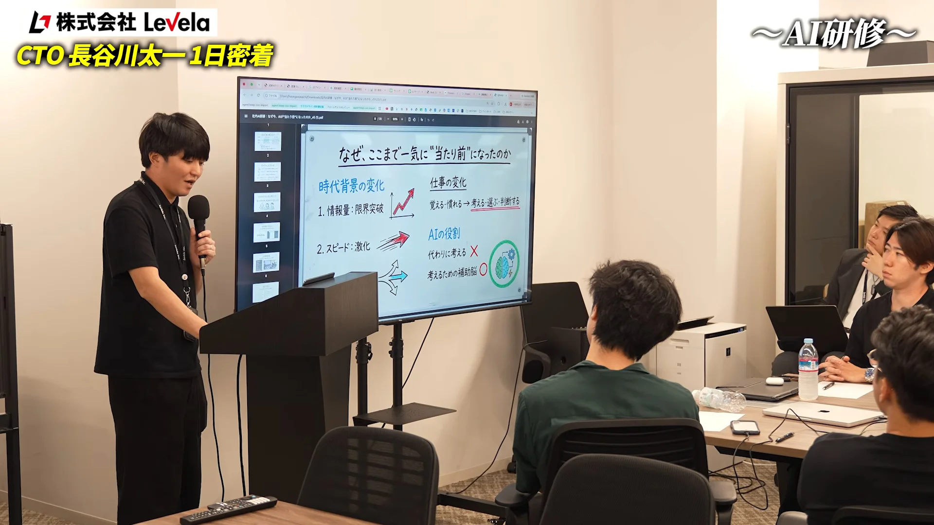 動画の字幕と大型モニターの研修スライド。演台で話す長谷川さんと、並んで座る参加者