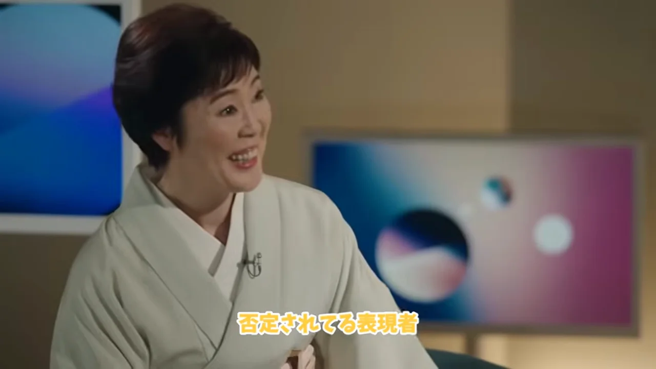 動画キャプチャ: テロップ「否定されてる表現者」が出て、聞き手が笑っている場面（1:04頃）