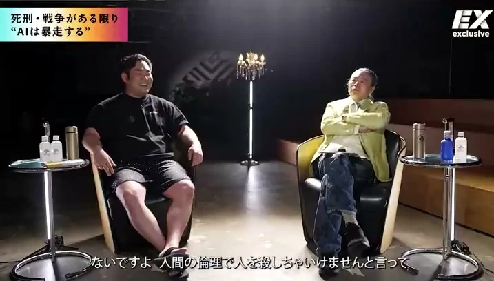 動画キャプチャ: NewsPicks対談の画面。「死刑・戦争がある限り “AIは暴走する”」のテロップが出て、2人が椅子に座って話している（8:27頃）